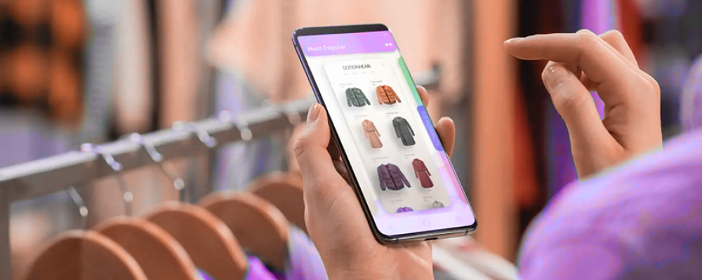 Pessoa usando smartphone para visualizar opções de roupas em um aplicativo, em frente a um cabideiro em uma loja física