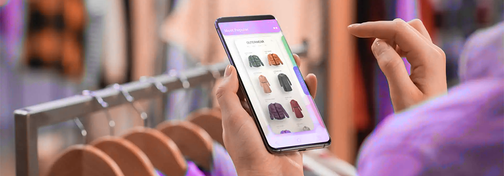 Pessoa usando smartphone para visualizar opções de roupas em um aplicativo, em frente a um cabideiro em uma loja física